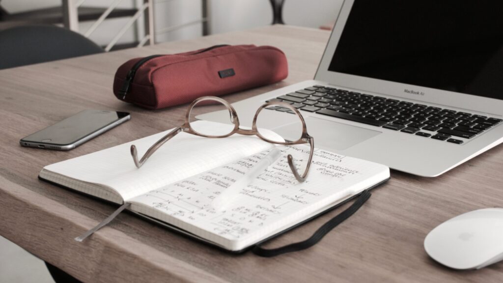 Notizbuch, auf dem eine Brille liegt, davor ein Federmäppchen. Alles vor einem aufgeklappten Laptop.
